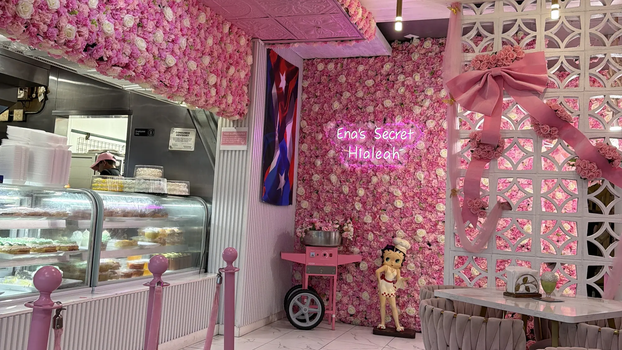 Sal&oacute;n de Ena&rsquo;s Secret Bake: pared de flores rosadas con un r&oacute;tulo de ne&oacute;n, vitrina de dulces, cordones rosados y sillas de terciopelo sobre suelo de m&aacute;rmol