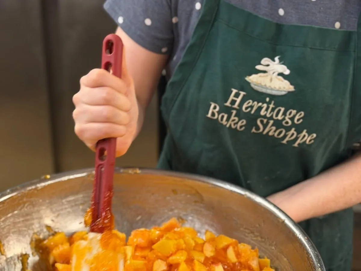 A baker in a Heritage Bake Shoppe apron stirring a large pan of cut peaches