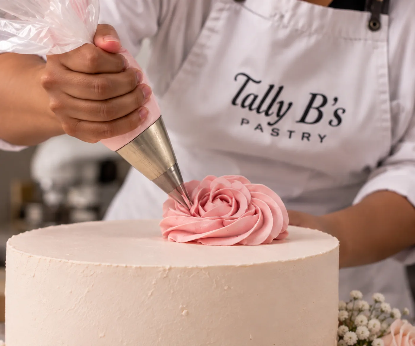Tally piping a pink buttercream rose onto a cake, wearing her Tally B's Pastry apron